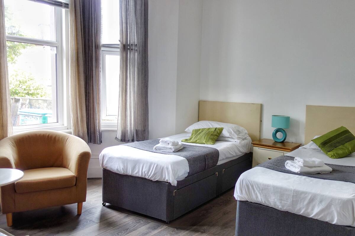 A hotel room with two beds, a chair, a nightstand with a lamp, and a window with curtains.