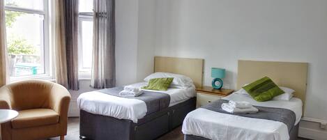 Habitación de hotel con dos camas, una silla, una mesita de noche con lámpara y una ventana con cortinas.