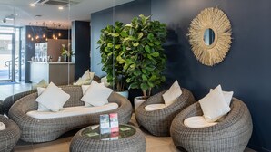 Un área de descanso moderna con sofás y otomanas de mimbre, una planta verde grande y un espejo decorativo en la pared.