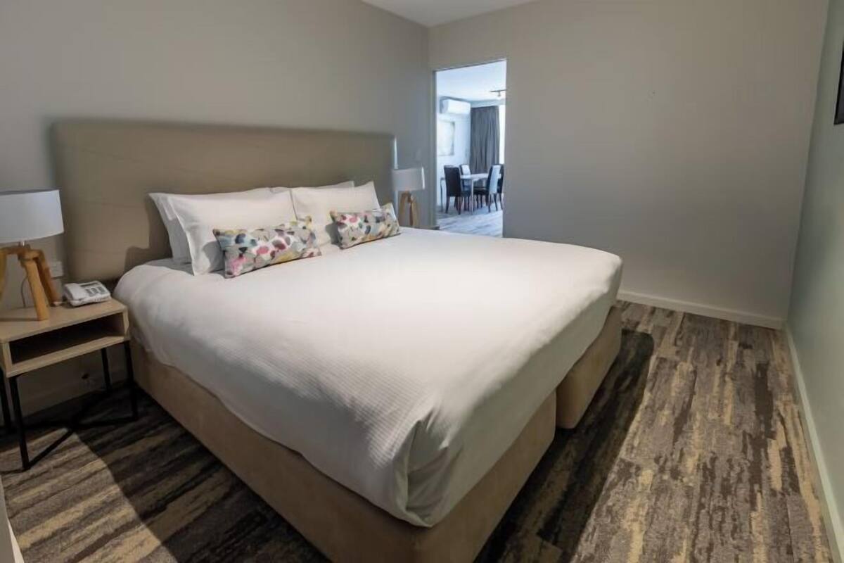 Habitación de hotel con una cama grande, una mesita de noche con lámpara y vistas al comedor.