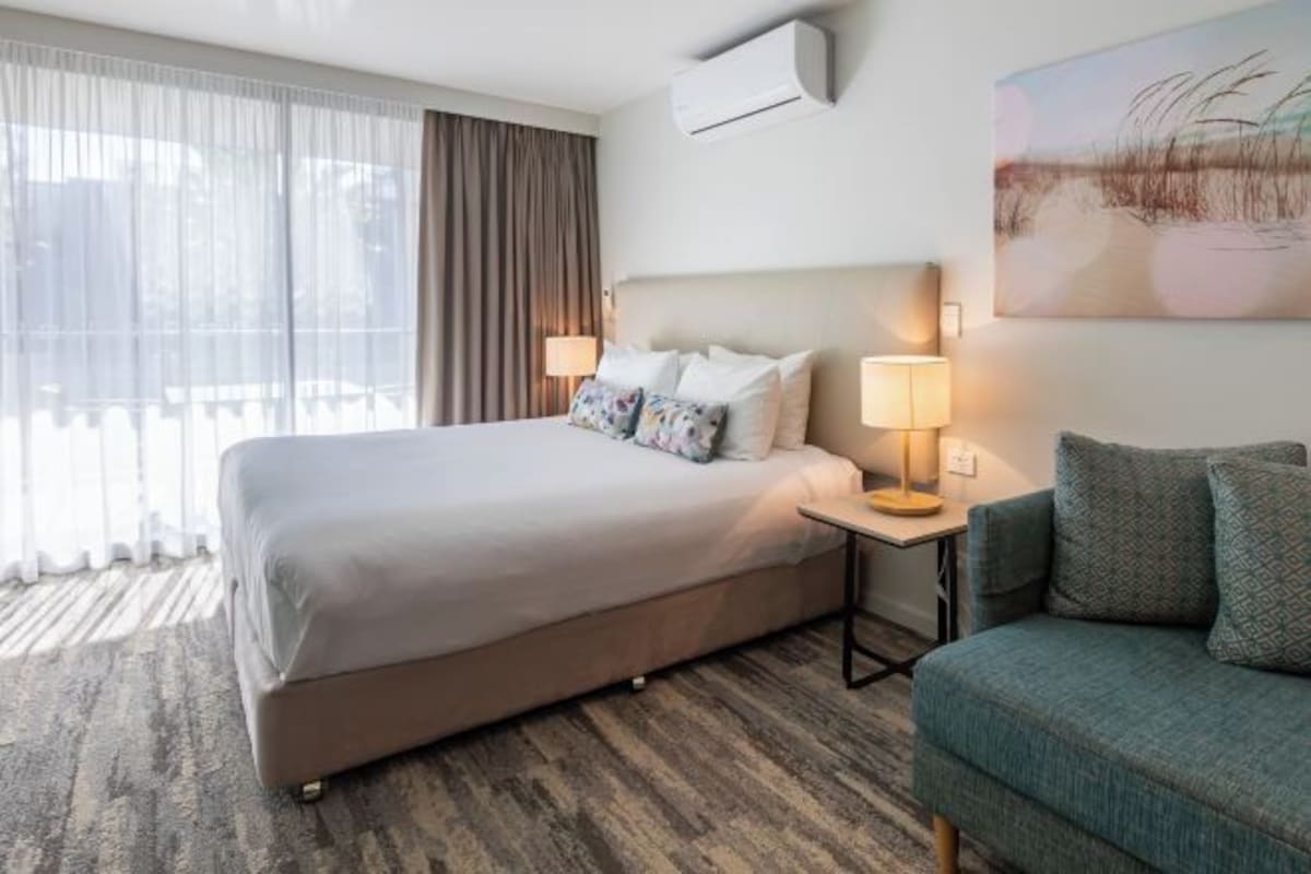Una habitación de hotel con una cama, una mesita de noche con lámpara, un sofá, un ventanal con cortinas y un equipo de aire acondicionado.