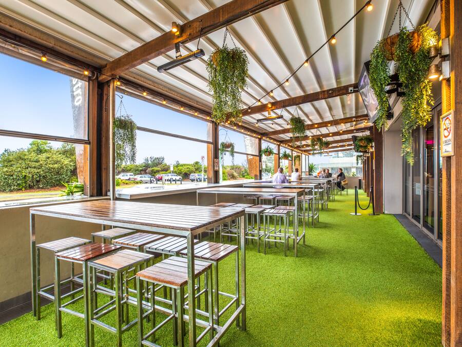Un restaurante con terraza al aire libre, césped artificial verde, plantas colgantes y vistas a la calle.