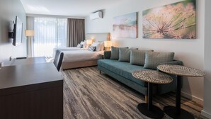 Una habitación de hotel con una cama, un sofá, dos mesitas auxiliares, un escritorio y un amplio ventanal con cortinas.