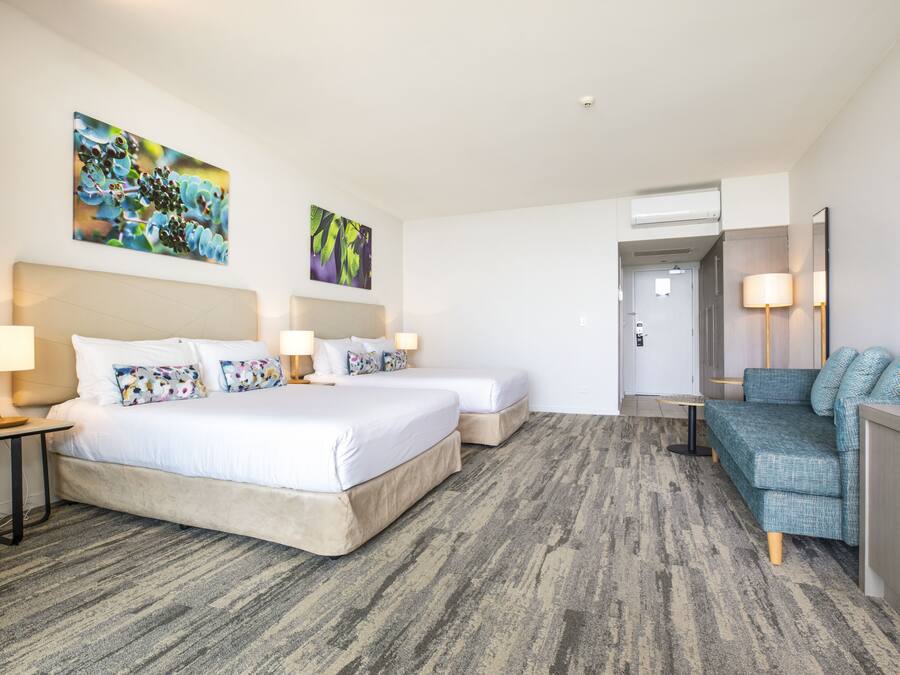 A hotel room with two beds, a sofa, and a desk. There are wall-mounted lamps and artwork above the beds.