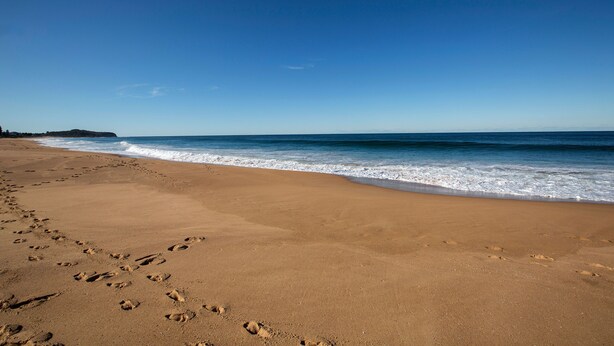 Una playa arenosa con huellas que conducen al agua, cielo despejado y una línea de costa distante.