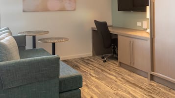 Un salon moderne comprenant un canapé d’angle, deux petites tables rondes, un téléviseur à écran plat fixé au mur et un coin cuisine avec des placards et un réfrigérateur.