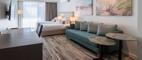 A hotel room with a bed, a sofa, two side tables, a desk, and a large window with curtains.