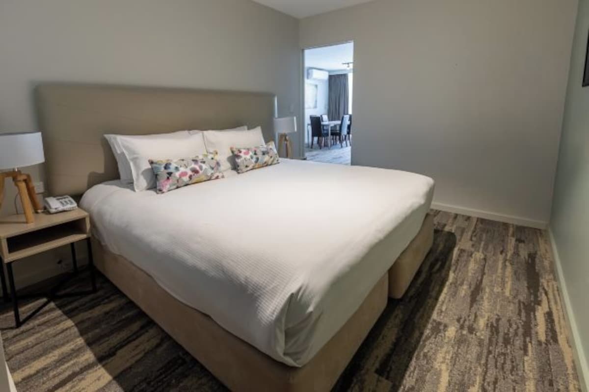 Habitación de hotel con una cama grande, una mesita de noche con lámpara y vistas al comedor.