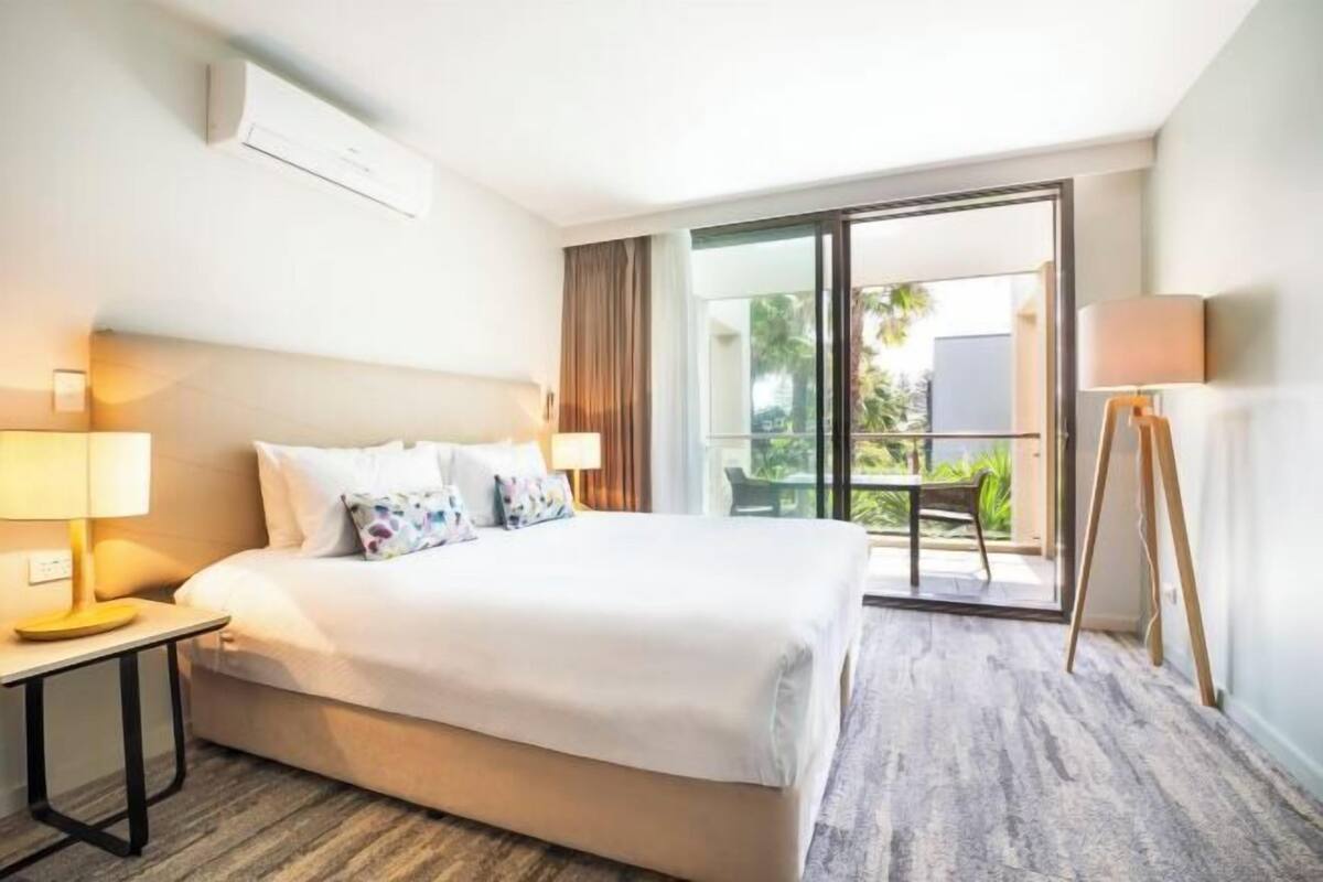 Habitación de hotel con cama, lámparas de noche, vistas al exterior y un balcón.