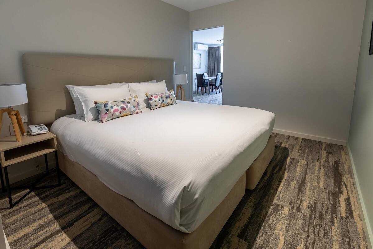 Habitación de hotel con una cama grande, una mesita de noche con lámpara y un teléfono. El suelo está alfombrado y se ve un comedor a través de una puerta abierta.