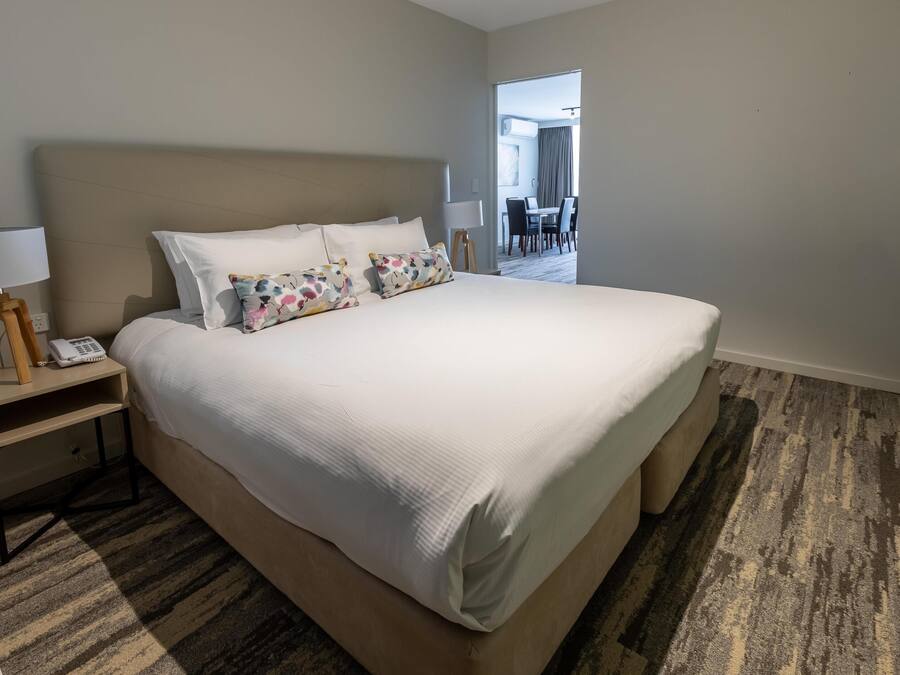 A hotel room with a large bed, a bedside table with a lamp, and a telephone. There is a carpeted floor and a view of a dining area through an open door.