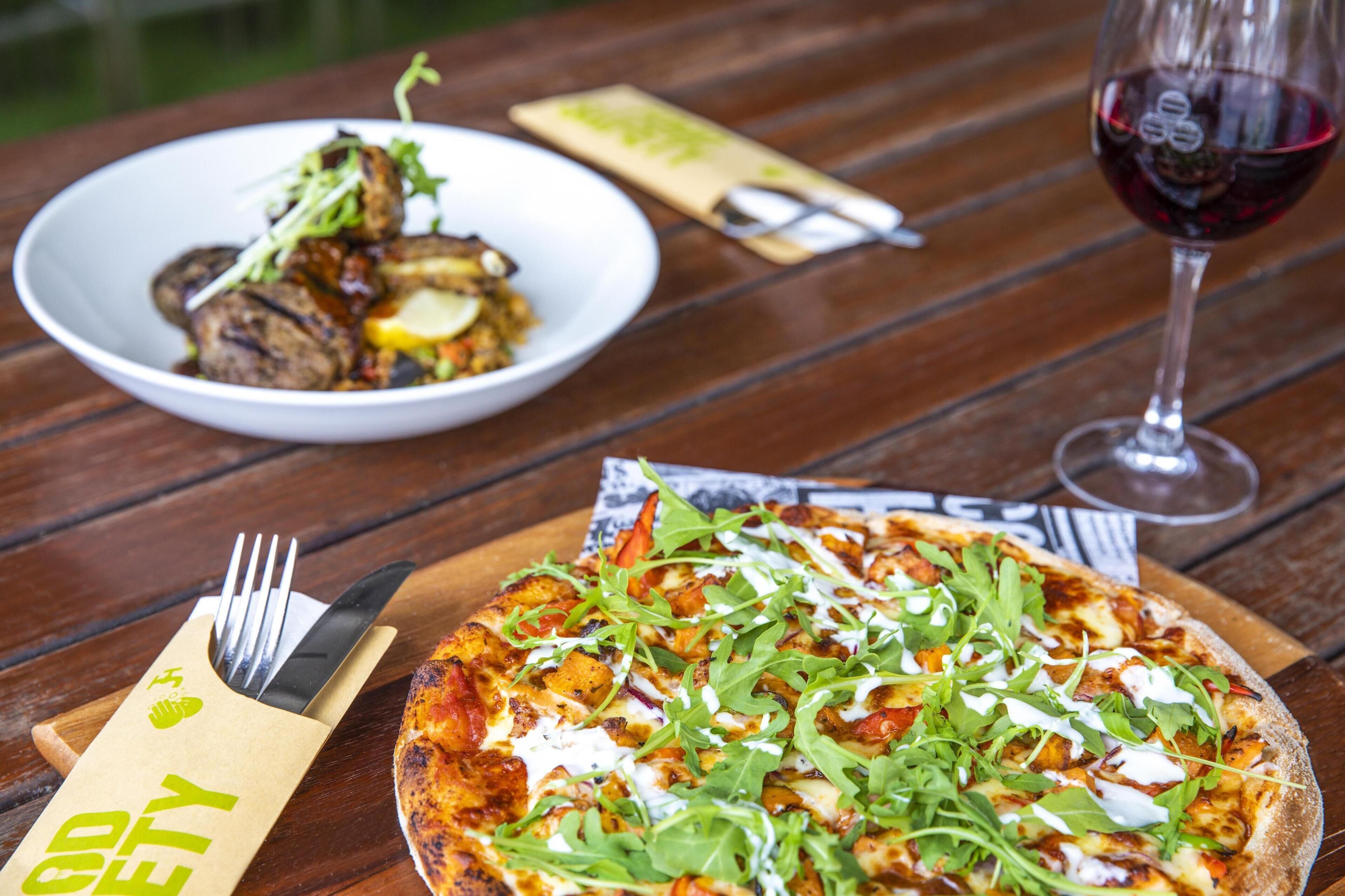 A pizza with greens and a bowl of food on a wooden table.