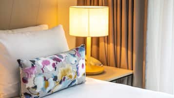 En pænt redt seng med en hvid pude og en farverig blomsterpude. En lampe med en beige skærm står på et natbord, og gardiner er halvt trukket til siden.