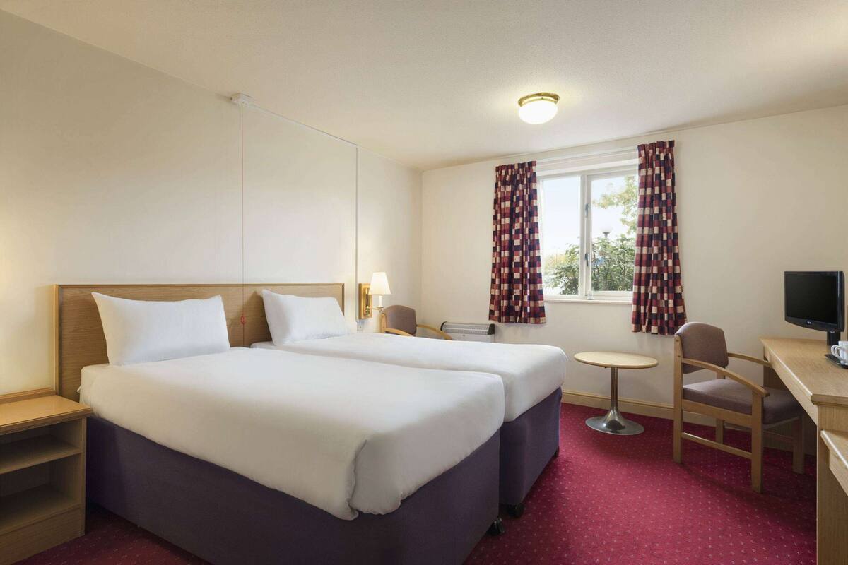 Une chambre d’hôtel avec deux lits, un bureau, une chaise, une télévision et une fenêtre avec des rideaux.