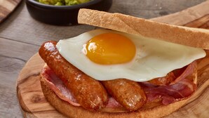 Un plato de desayuno con un huevo frito, salchichas y tocino, sobre una tabla de madera.