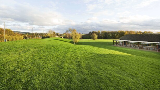Un campo cubierto de césped con un edificio que tiene asientos al aire libre y una zona techada.
