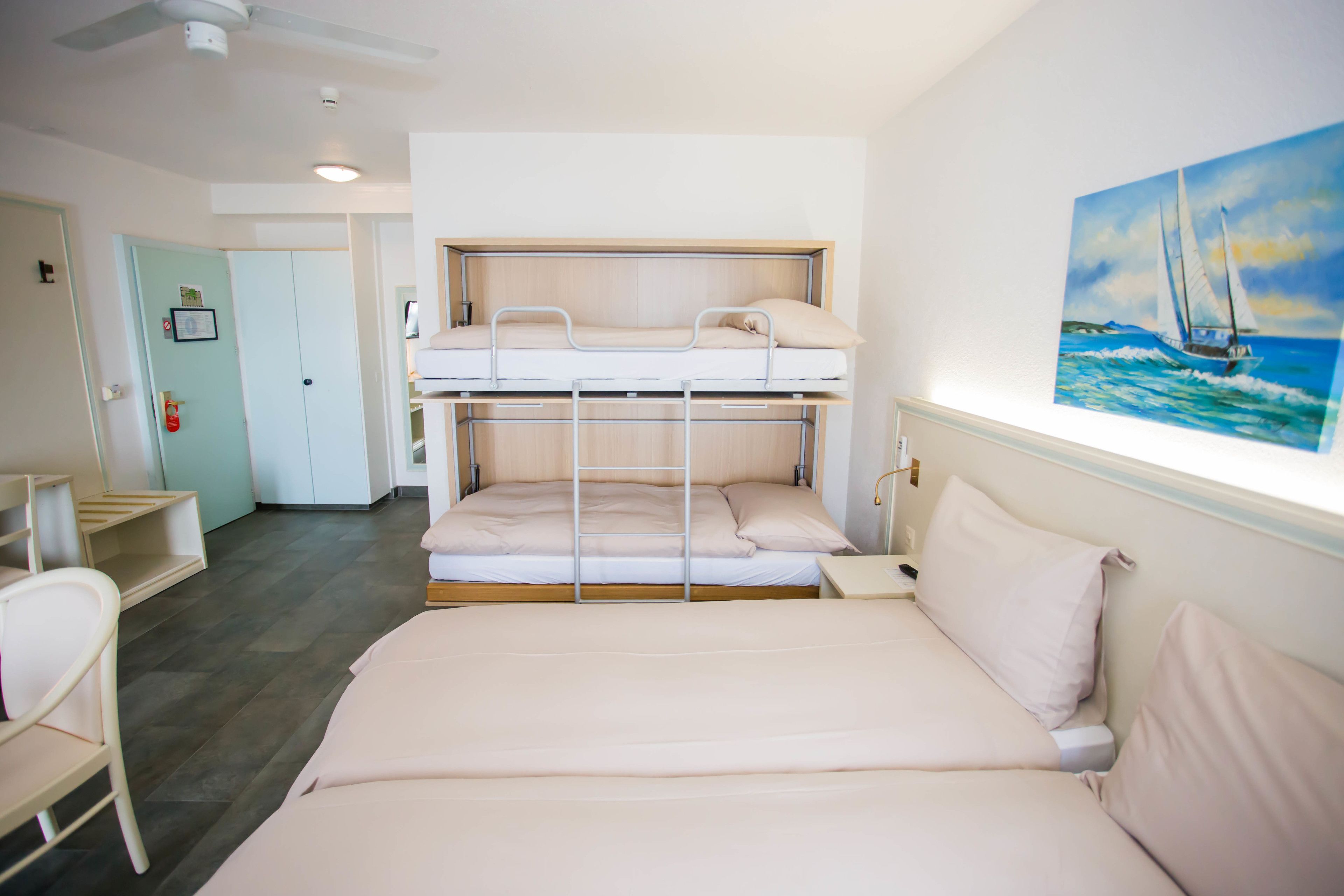 Een kamer met een stapelbed, een bureau en een stoel. Er hangt een schilderij aan de muur en een deur met een handvat.