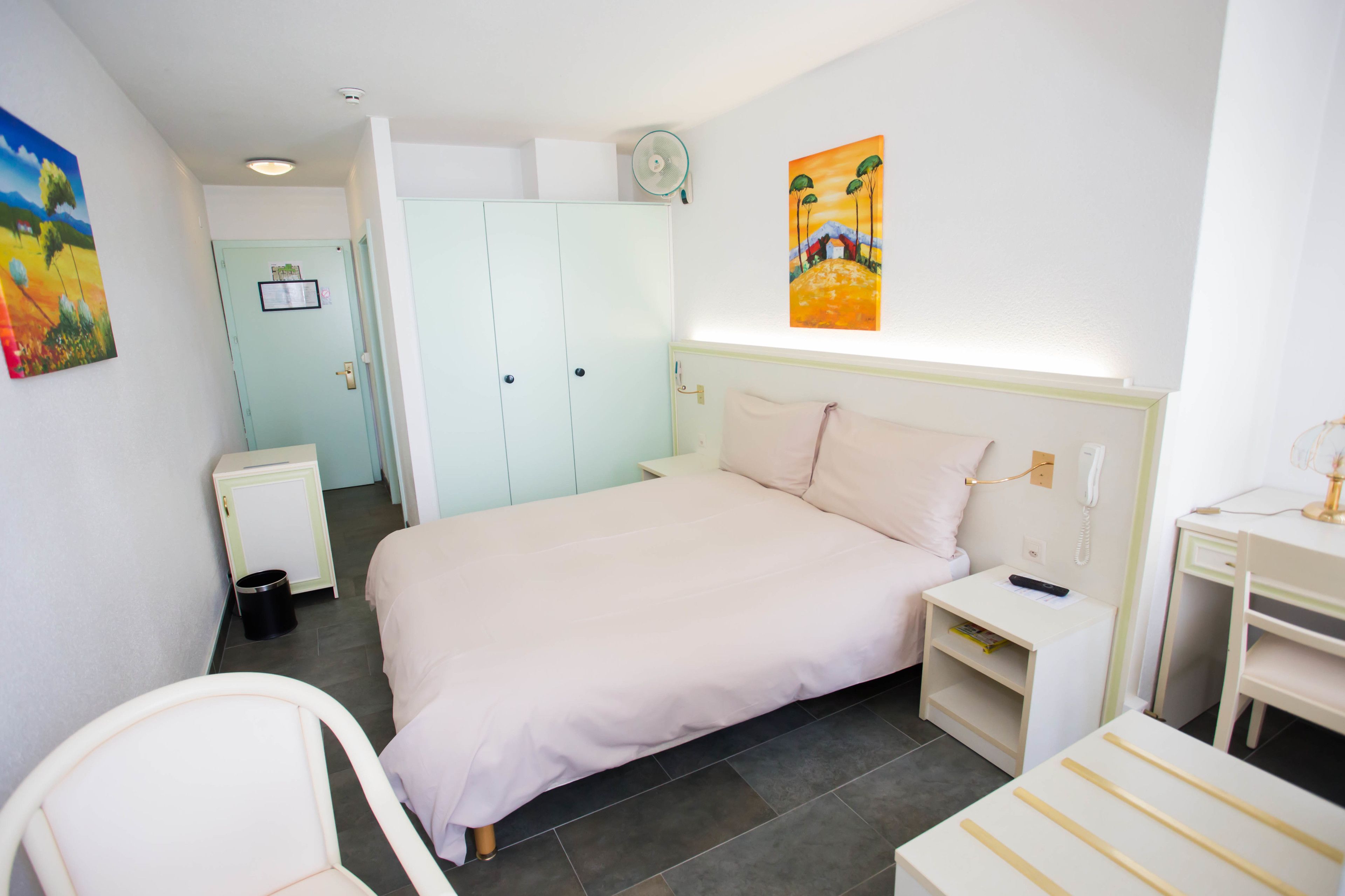 A bedroom with a bed, bedside tables, a desk, a chair, a wardrobe, and a painting on the wall.