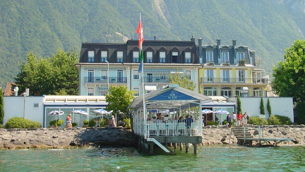 A waterfront hotel with a dock, outdoor seating area, and a mountain backdrop. - Hotel Du Port (Villeneuve)