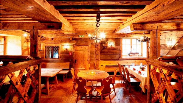 Una cabaña rústica de madera con una mesa central rodeada de sillas, paredes de madera y techo inclinado con vigas a la vista.