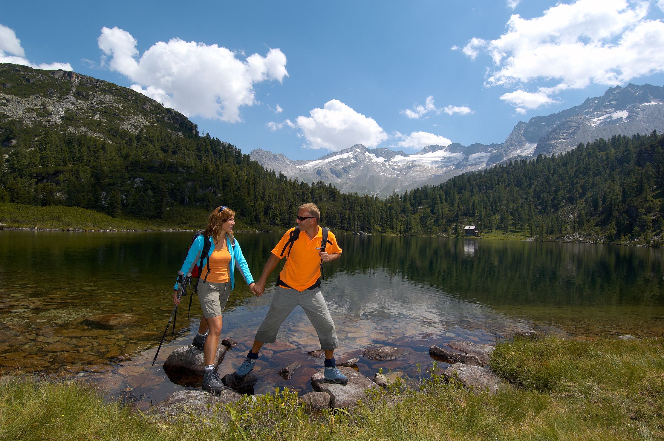 Two hikers on a rocky shore by a lake with mountains in the background.