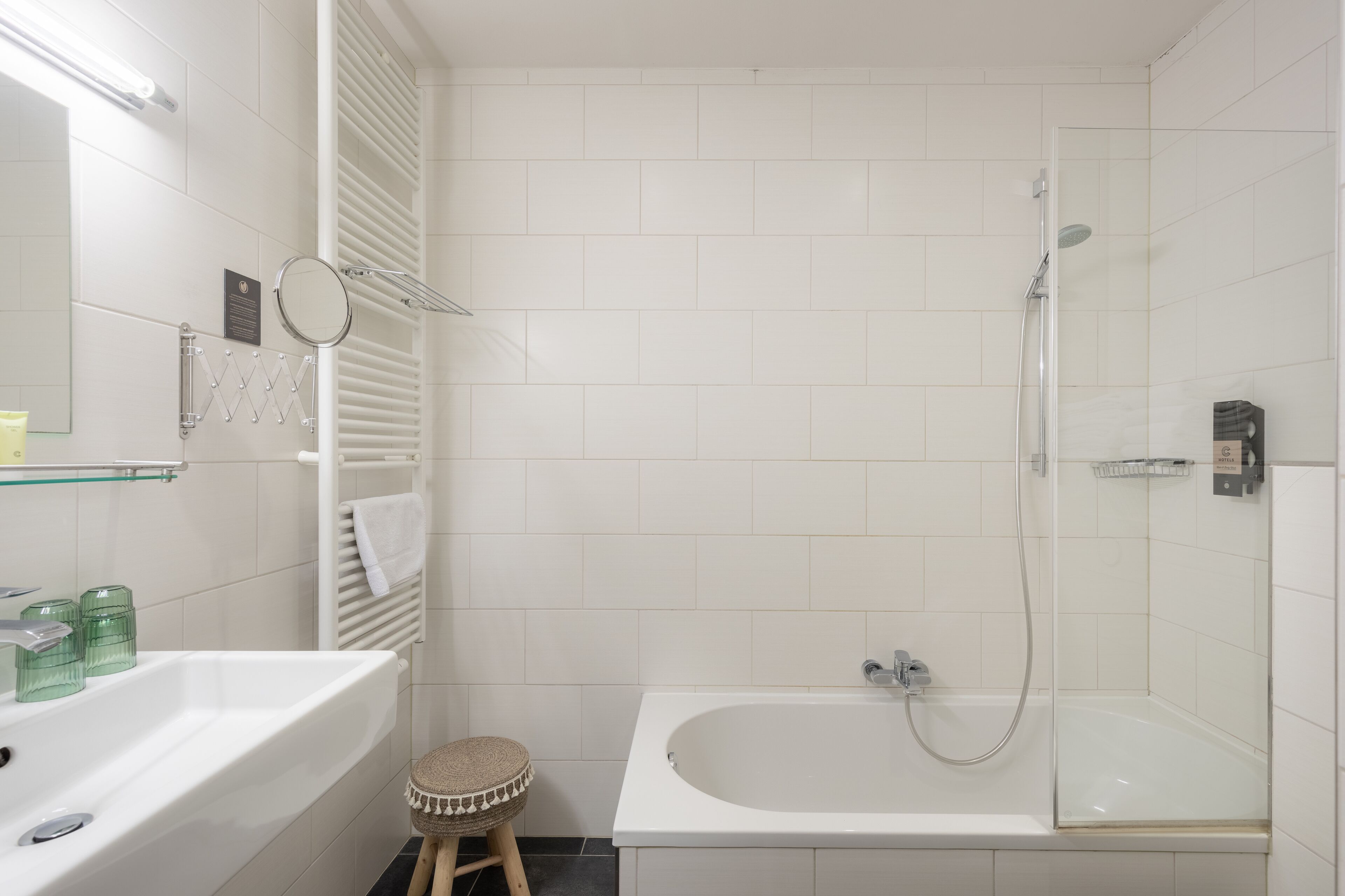 Une salle de bain moderne avec un lavabo blanc, un miroir rond, une étagère en verre, un porte-serviettes, un petit tabouret et une baignoire équipée d’un pommeau de douche.