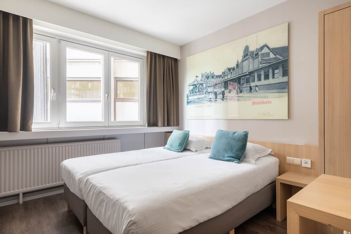 Quarto de hotel com duas camas, uma janela com cortinas, um radiador, uma mesa de madeira e um quadro pendurado na parede retratando um edifício.