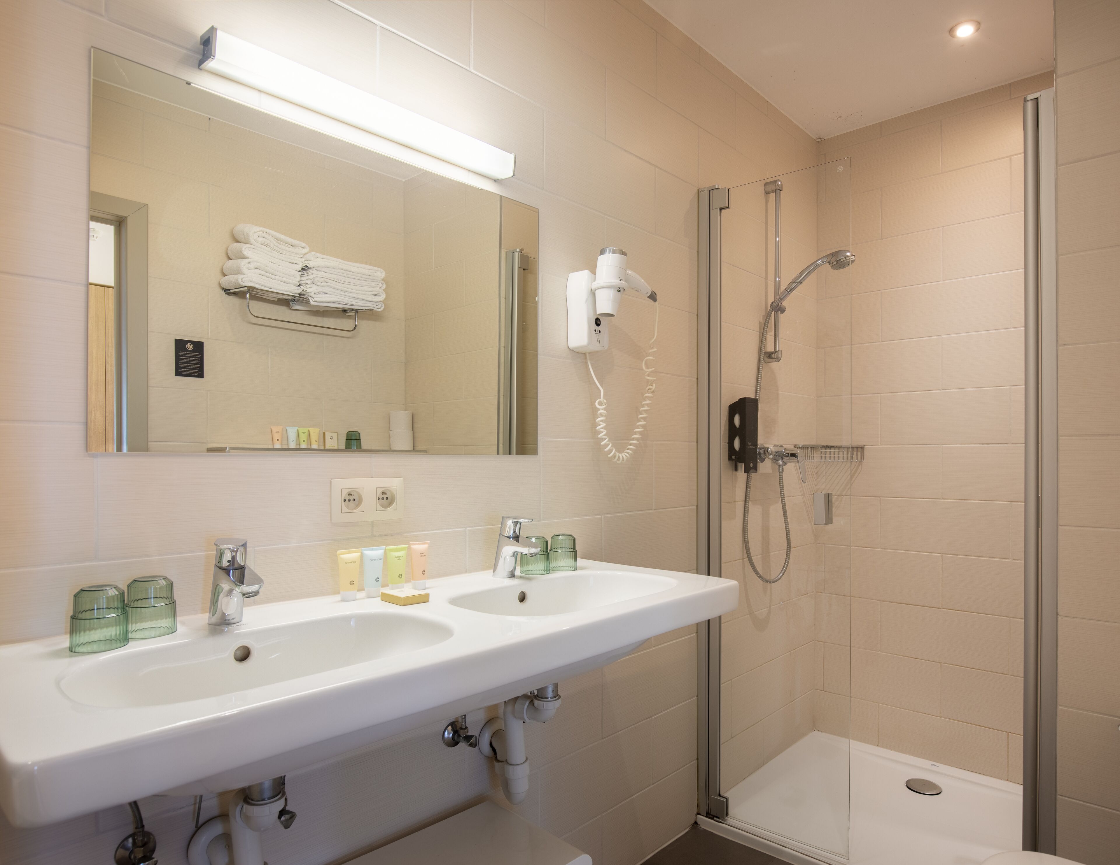 A bathroom with a white sink, two glass soap dispensers, a mirror, and a glass-enclosed shower area.