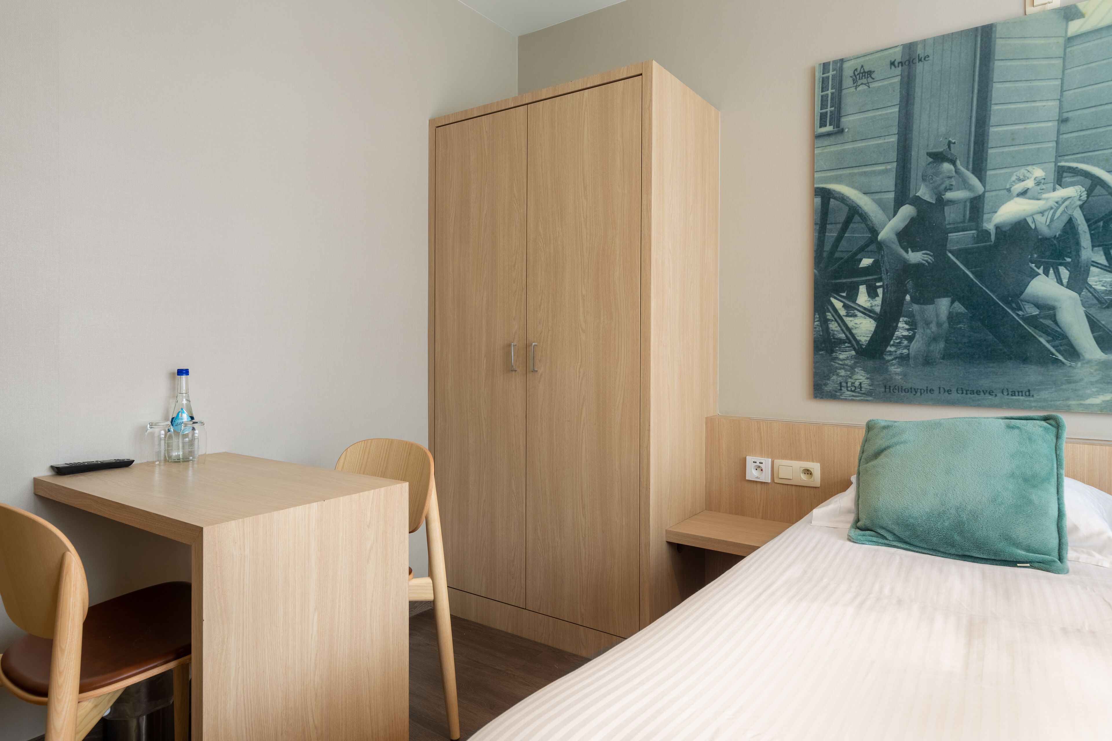 Una habitación de hotel con un armario de madera, un escritorio con silla, una cama con almohada y un cuadro colgado en la pared.