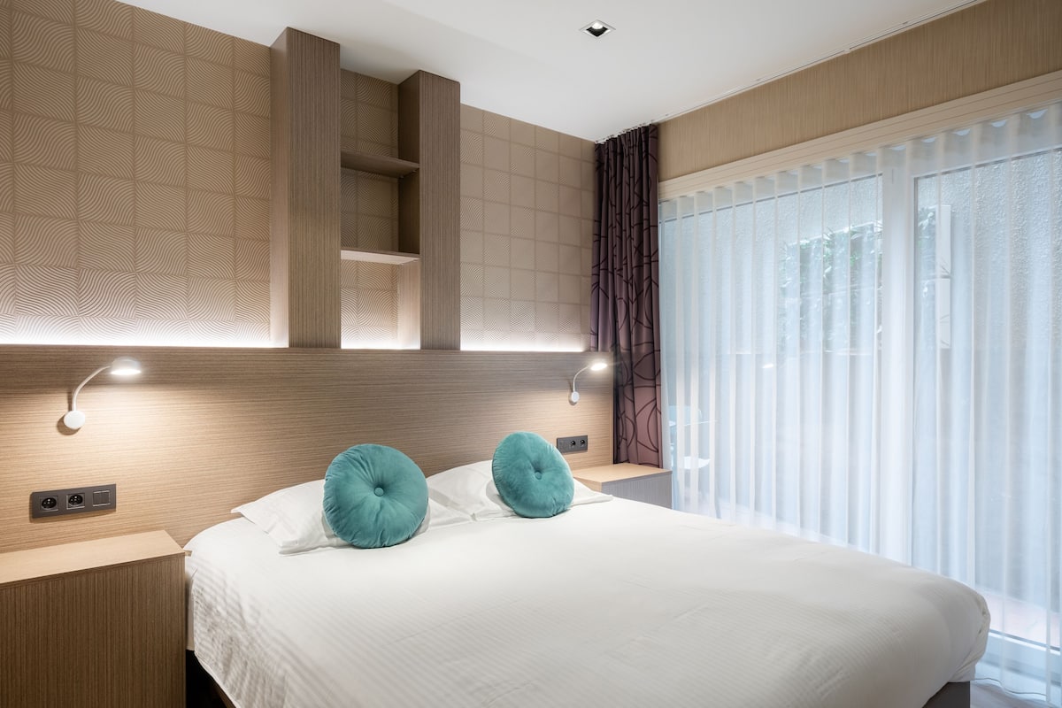 Quarto de hotel moderno com uma cama grande, duas almofadas verde-água, cabeceira de madeira, prateleiras embutidas e uma janela grande com cortinas leves.