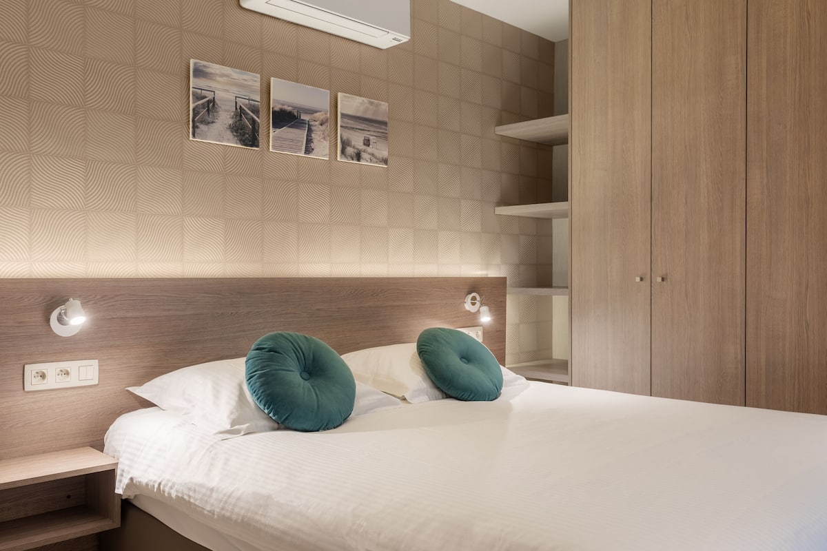 Quarto com cabeceira de madeira, cama com roupa de cama branca e almofadas verde-água, mesa de cabeceira e prateleira fixada na parede.