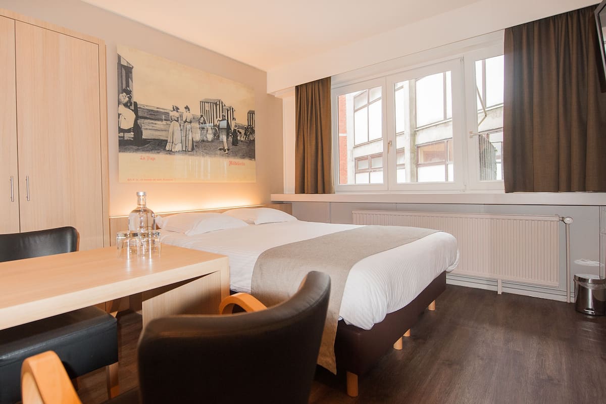 Quarto de hotel com uma cama, uma escrivaninha, uma cadeira, um quadro na parede e uma janela com cortinas.