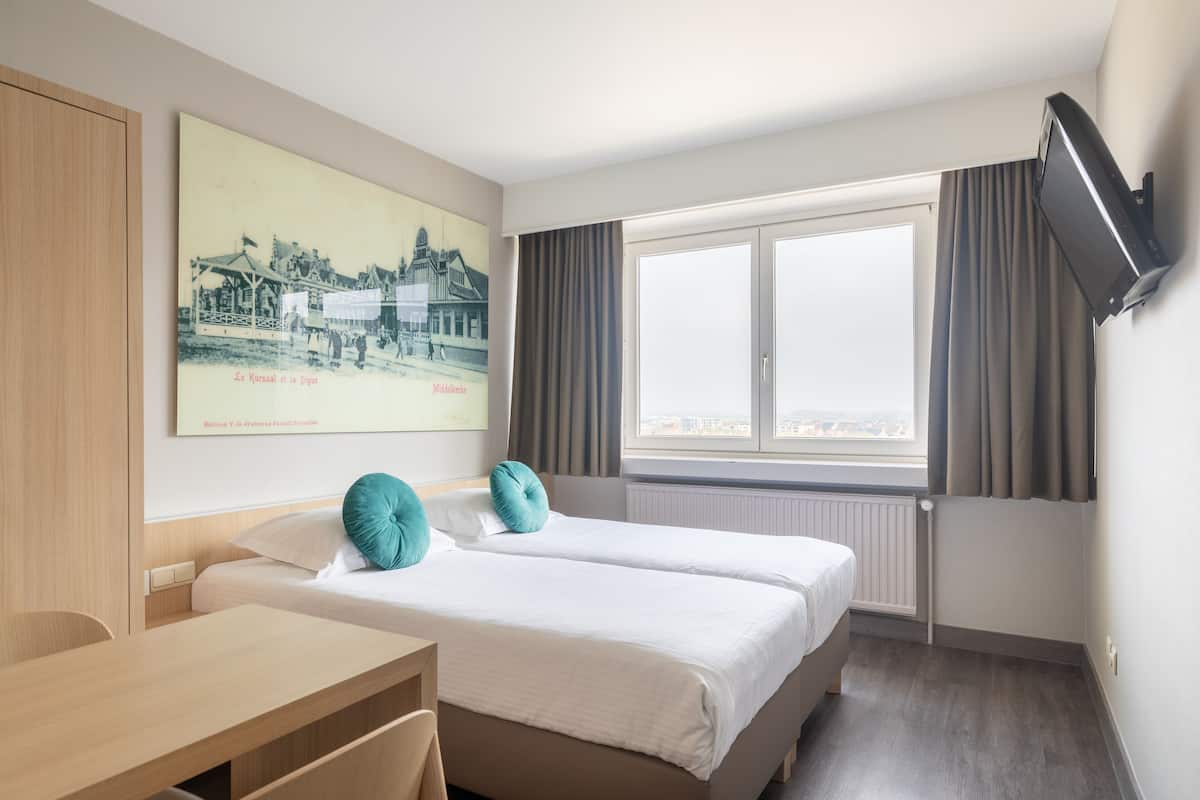Quarto de hotel com duas camas, uma escrivaninha, televisão presa na parede e uma janela grande com cortinas.