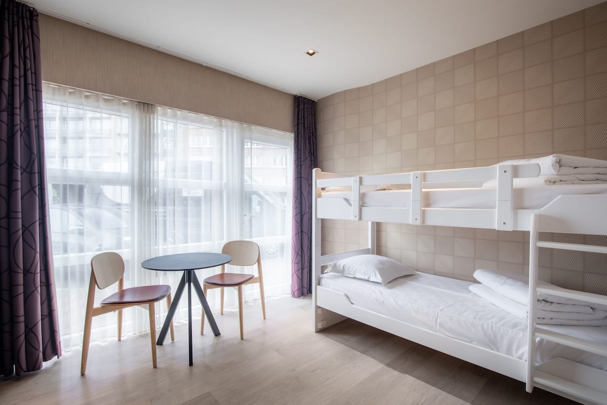 Quarto de hotel com beliche, mesa redonda, duas cadeiras e janelas amplas com cortinas leves.