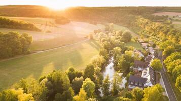 A rural landscape with a house surrounded by trees and fields, a river, and a road.