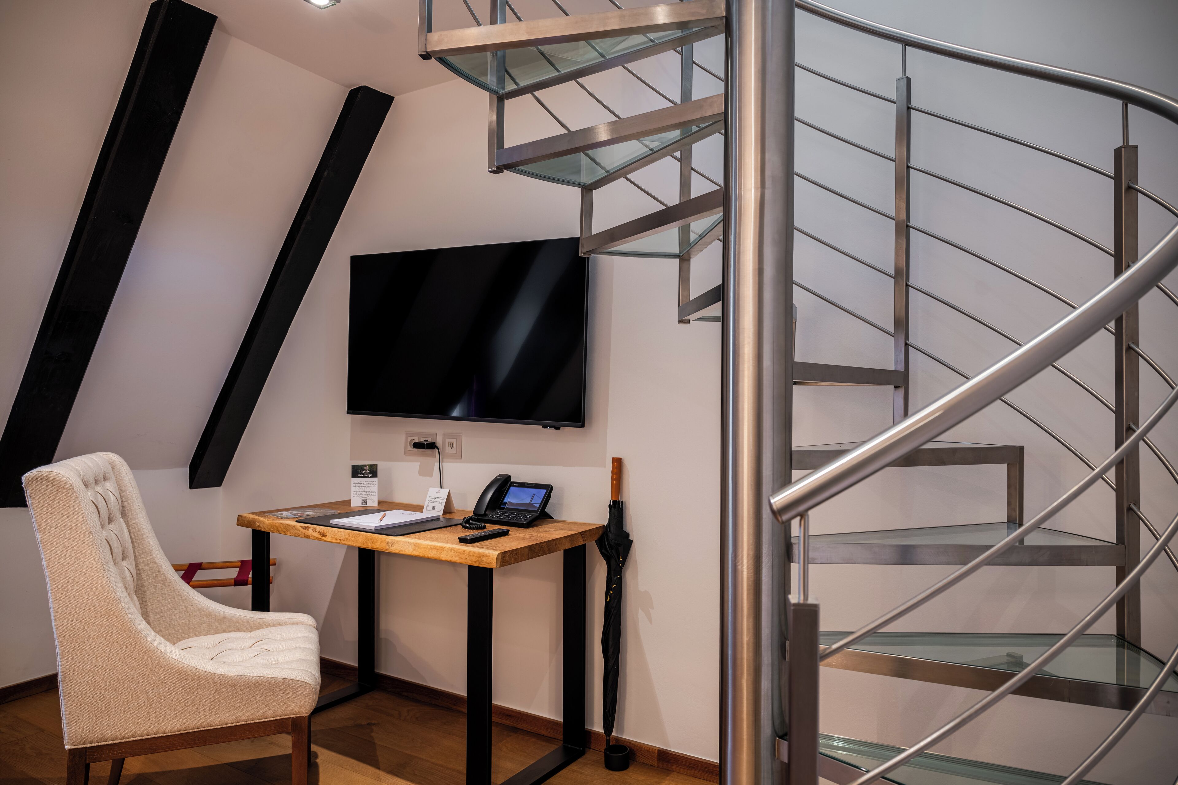 Ein modernes Zimmer mit Schreibtisch aus Holz, einem Sessel, einem Flachbildfernseher und einer Wendeltreppe.