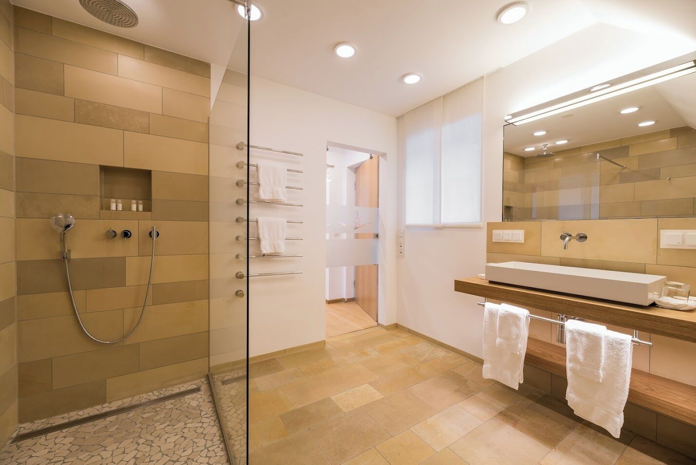 Un baño moderno con cabina de ducha de vidrio, tocador de madera con un espejo grande y piso de baldosas.