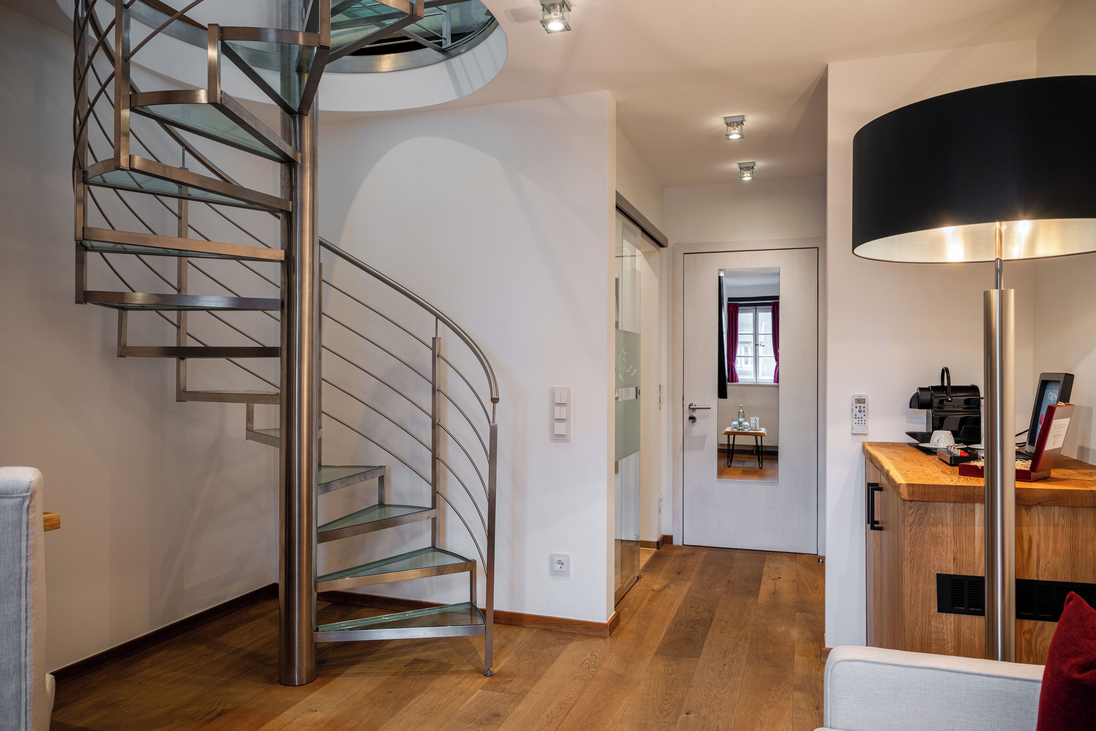 A modern interior with a spiral staircase, a wooden floor, a desk with a lamp, and a mirror reflecting a room with a window.
