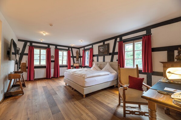 Un dormitorio con una cama grande, una silla de madera, una mesa con una lámpara y una ventana con cortinas rojas.