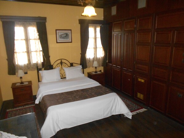 A neatly made bed with white linens, a wooden headboard, and a nightstand with a lamp. There is a large wooden wardrobe to the right and a framed picture on the wall above the bed.