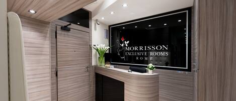 A reception desk with a modern design, a large window displaying the name "MORRISON EXCLUSIVE ROOMS ROME," and a potted plant on the counter.