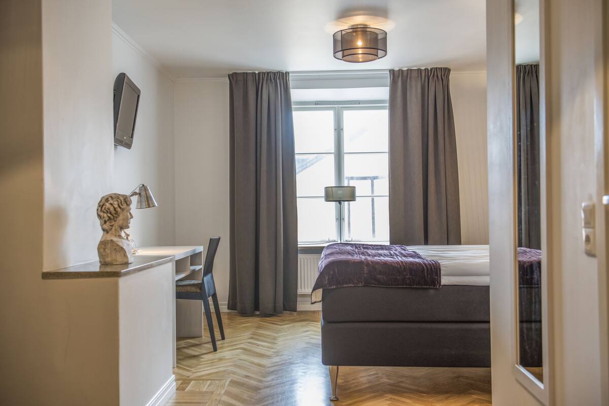 A bedroom with a bed, a desk, and a chair. There is a window with curtains, a lamp, and a sculpture on the wall.