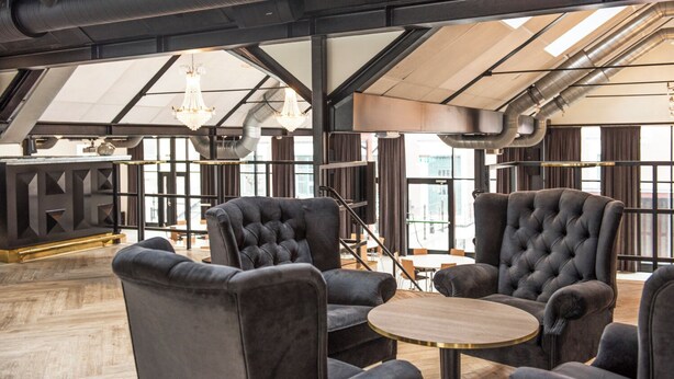 A modern interior with high ceilings, exposed ductwork, and large windows. There are plush armchairs and a round table in the foreground. - Donners Hotel (Visby)