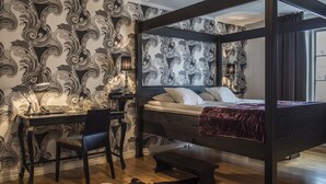 A four-poster bed with a dark headboard, a desk with a chair, and a patterned wallpaper. - Donners Hotel (Visby)