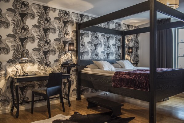 A four-poster bed with a dark headboard, a desk with a chair, and a patterned wallpaper. - Donners Hotel (Visby)