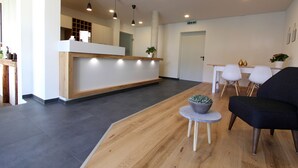 A reception area with a wooden reception desk, a small round table with a plant, and a black sofa. - Adler Hotel & Gasthaus (Nonnenhorn)