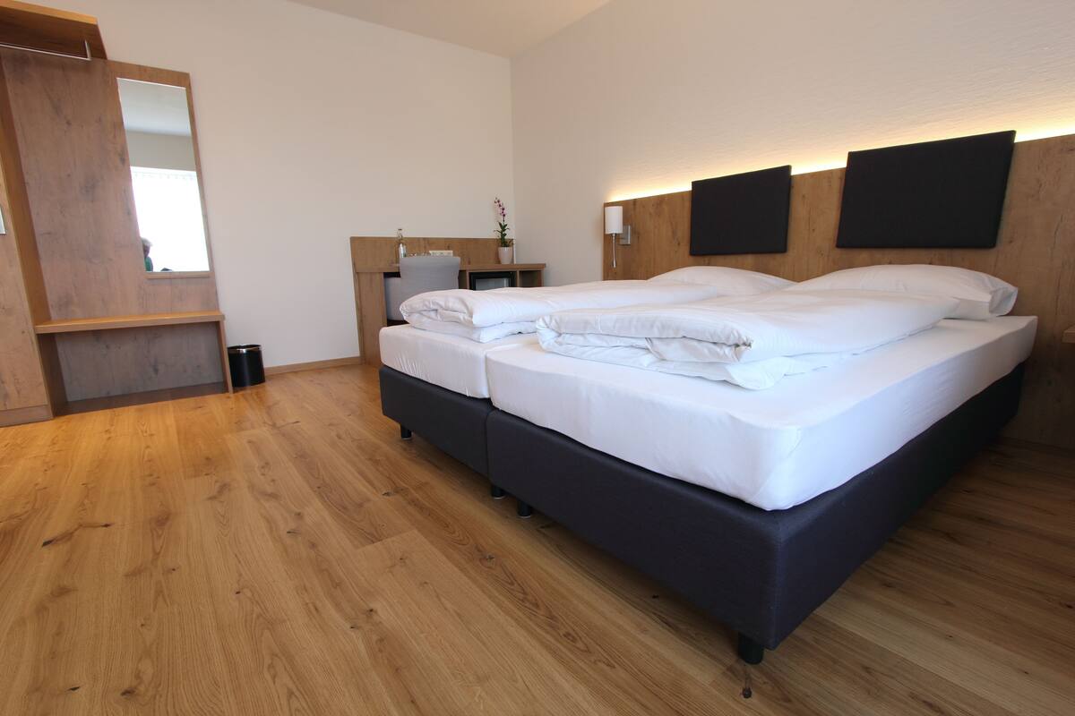 A modern hotel room with a large bed, wooden flooring, a small desk with a chair, and a mirror.