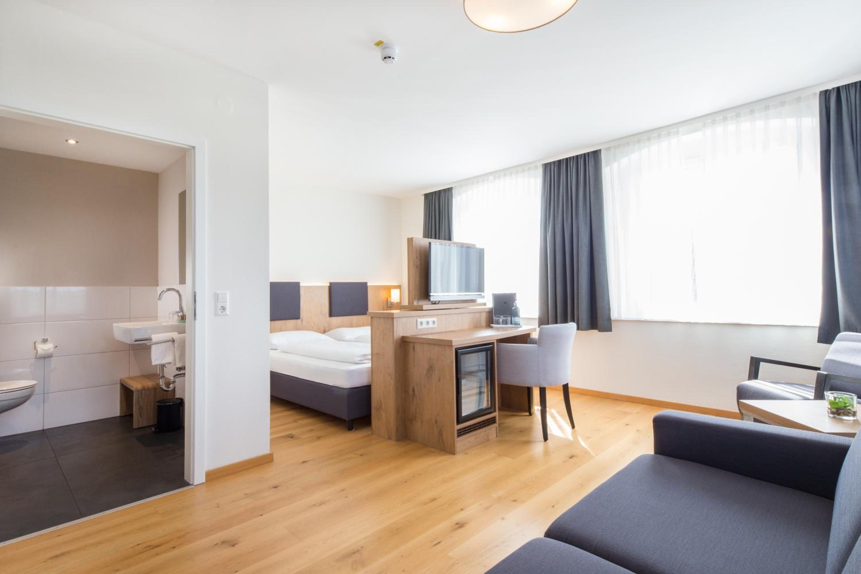Ein modernes Hotelzimmer mit Holzboden, einem Bett, einem Schreibtisch mit Computer, einem Fernseher und einer Sitzecke mit Sofa und Sesseln.