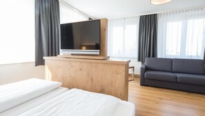 A modern hotel room with a large bed, a flat-screen TV on a wooden console, a gray sofa, and large windows with sheer curtains. - Adler Hotel & Gasthaus (Nonnenhorn)