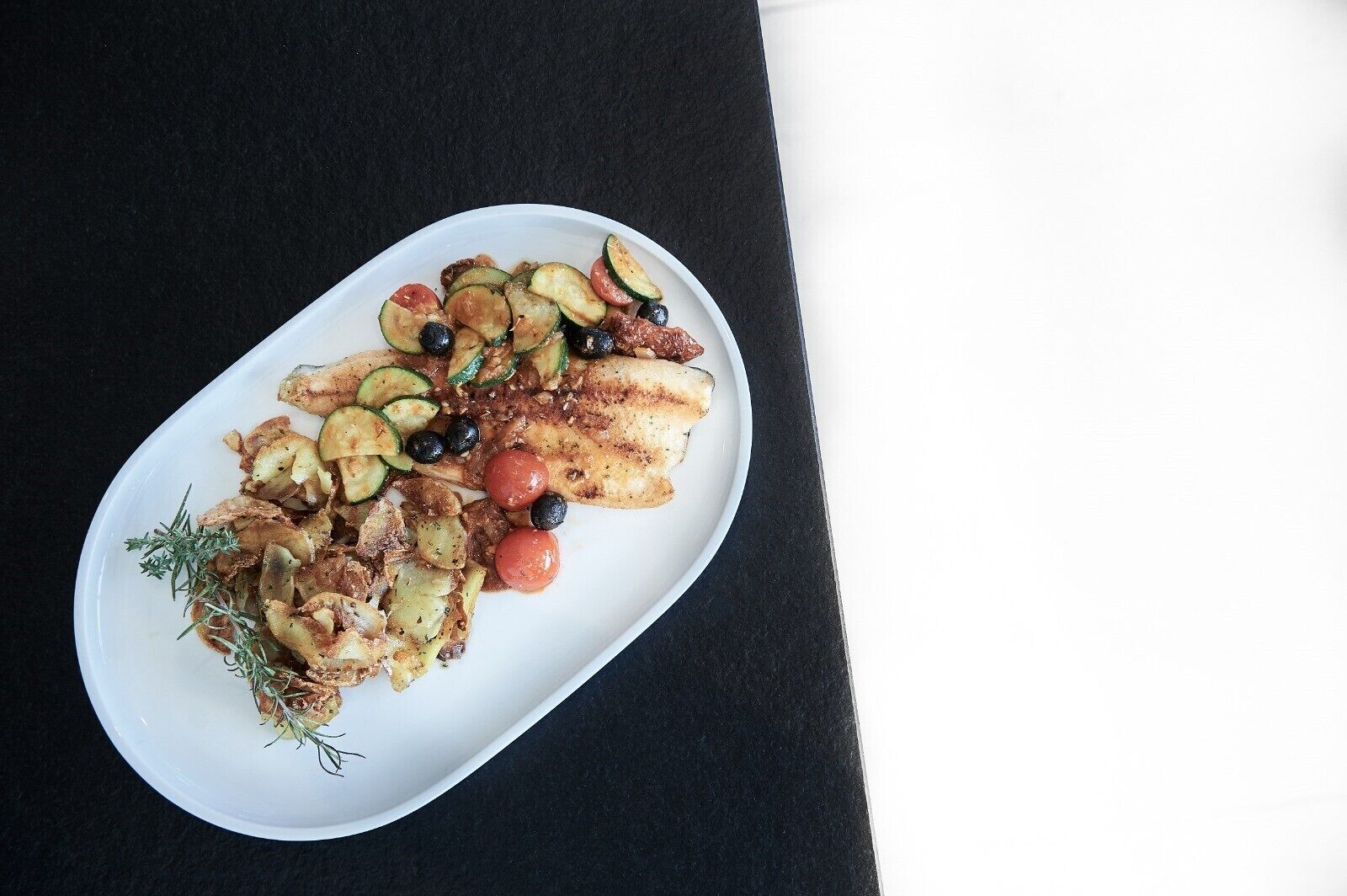 Une assiette ovale blanche contenant un plat composé de poisson grillé, de pommes de terre rôties, de rondelles de courgettes, de tomates cerises et d’olives noires, le tout agrémenté d’herbes aromatiques.